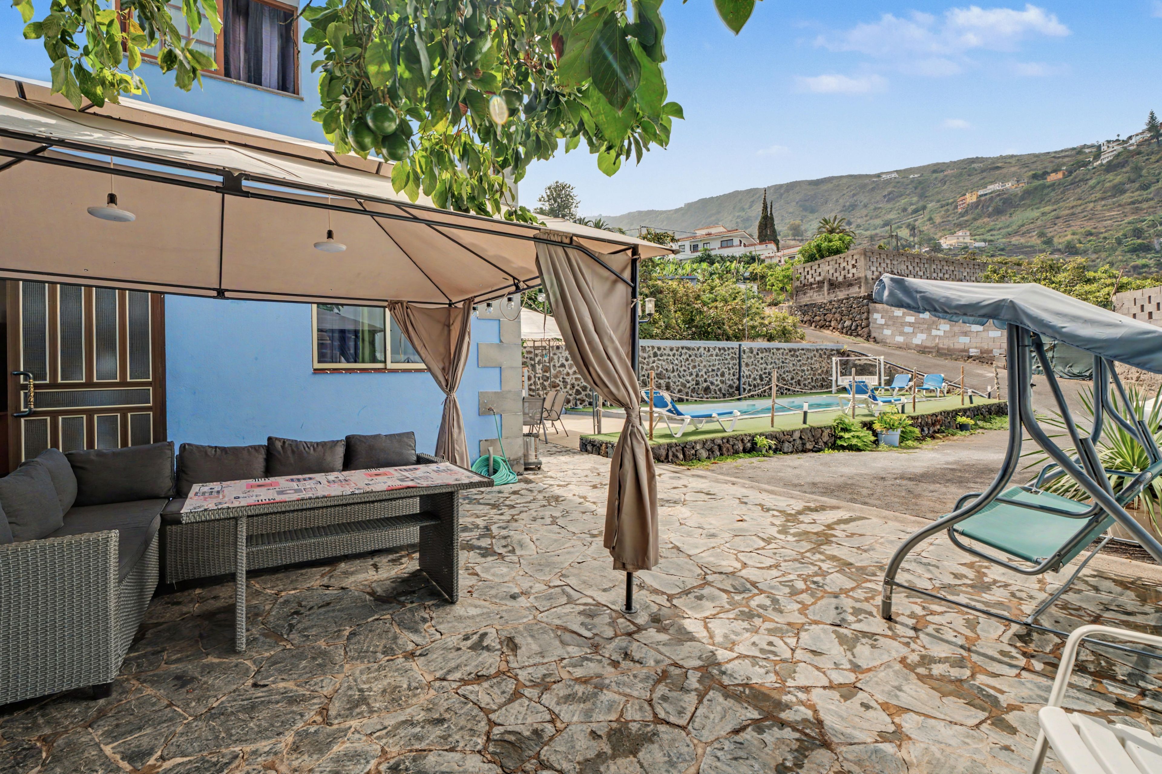 A patio area with a stone floor, outdoor furniture, a covered seating area, and a view of a pool and hillside.