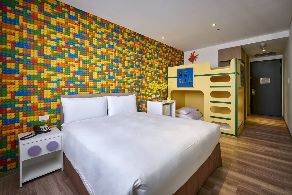 A bedroom with a colorful LEGO wall, a bed, a nightstand, and a small desk with a vase of flowers.