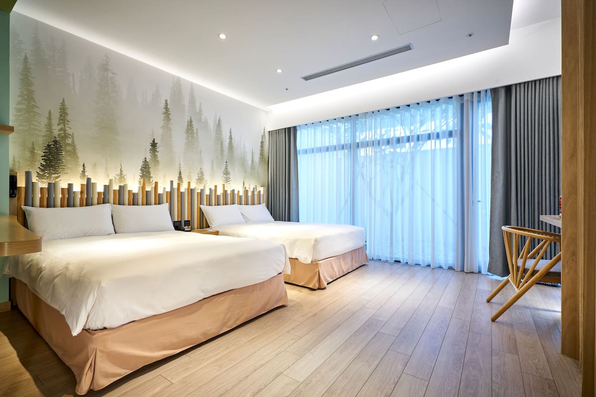 A modern hotel room with two beds, a large window with sheer curtains, a wooden floor, and a wall mural of trees.