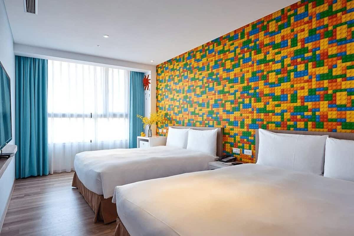 A hotel room with two beds, a colorful LEGO wall, a TV, and a window with curtains.
