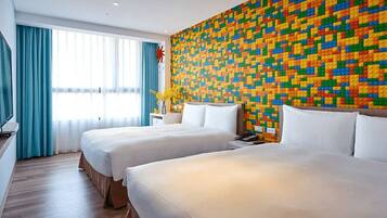 Habitación de hotel con dos camas, una pared decorada con LEGO de colores, televisor y ventana con cortinas.