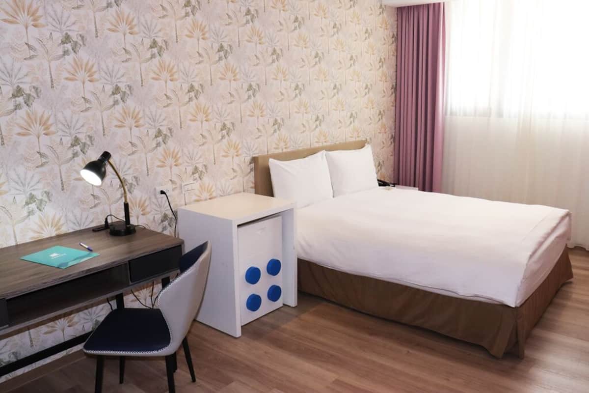 A hotel room with a bed, a desk with a lamp, a chair, and a small side table.