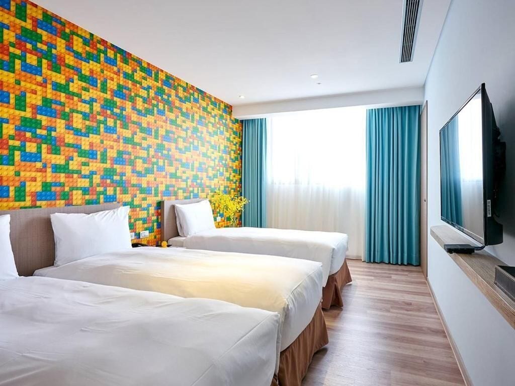 Hotellihuone, jossa on kaksi sänkyä, värikäs mosaiikkiseinä, televisio ja ikkuna, jossa on verhot.