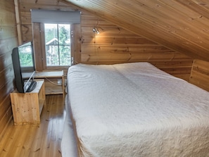 Un dormitorio con techo de madera, una cama con colcha blanca, un televisor sobre un mueble de madera y una ventana con vista a edificios.