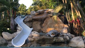 Una piscina con tobogán de agua, rocas y una estatua de cocodrilo.