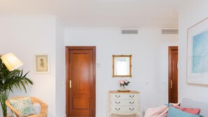 Una sala de estar con un sofá, un sillón de mimbre, una mesita con una lámpara, un aparador blanco y un espejo con marco en la pared.