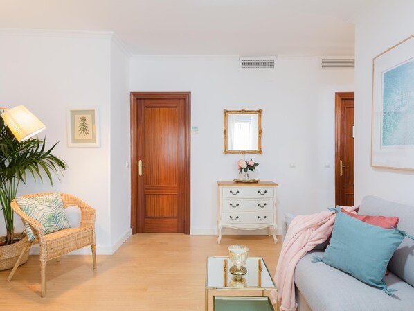 Una sala de estar con una silla de mimbre, un sofá, una mesita con superficie de vidrio, un aparador blanco con cajones y una puerta de madera.