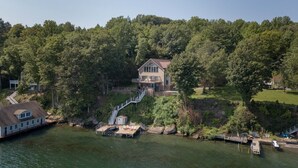 Una casa grande con una larga escalera que lleva a un muelle junto a un lago, rodeada de un bosque denso.