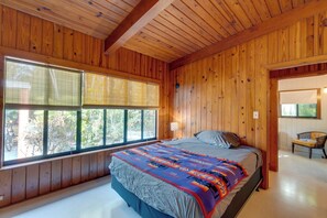 Un dormitorio con paredes de madera, un ventanal con persianas, una cama con manta colorida y una silla junto a la ventana.