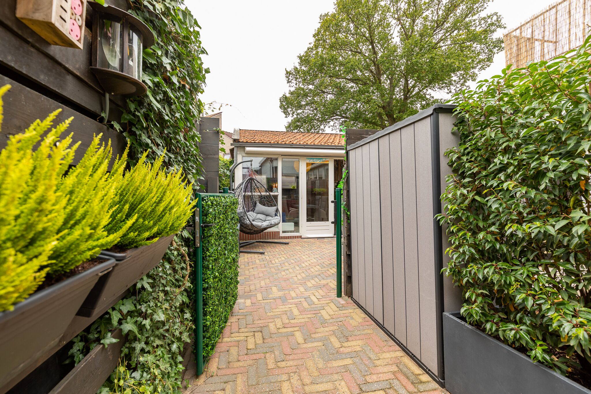 Un étroit sentier en briques mène à une maison moderne dotée d’un toit en tuiles, d’un fauteuil suspendu et d’un jardin planté de verdure.