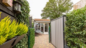 A narrow brick pathway leading to a modern house with a tiled roof, a hanging chair, and a garden with green plants.