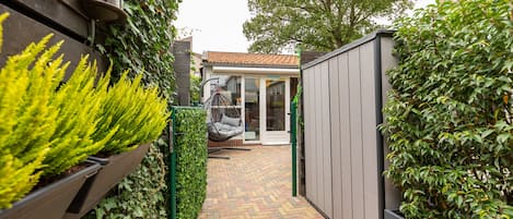 A narrow brick pathway leading to a modern house with a tiled roof, a hanging chair, and a garden with green plants.