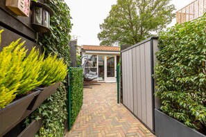 Uma estreita passagem de tijolos leva a uma casa moderna com telhado em telhas, uma cadeira suspensa e um jardim com plantas verdes.