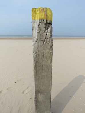 Um poste de madeira desgastado, com números e letras entalhadas, erguido sobre uma praia arenosa, com um céu claro ao fundo.