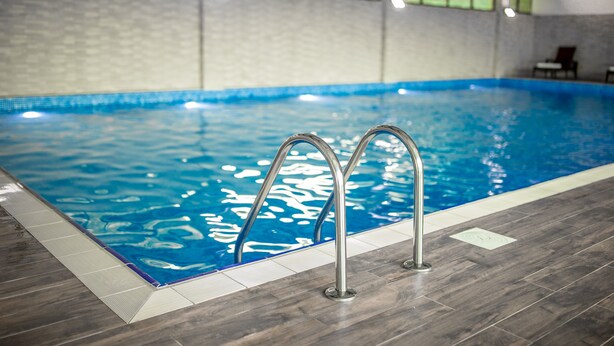 Una piscina interior con agua azul cristalina, escaleras de metal y suelo de baldosas.
