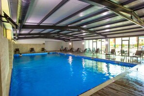 Una piscina interior con agua azul cristalina, rodeada de tumbonas y un techo alto con vigas a la vista.