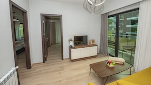 Una sala de estar moderna con piso de madera, un sofá, una mesa de centro con frutas, un televisor sobre un mueble bajo y un balcón con vistas.