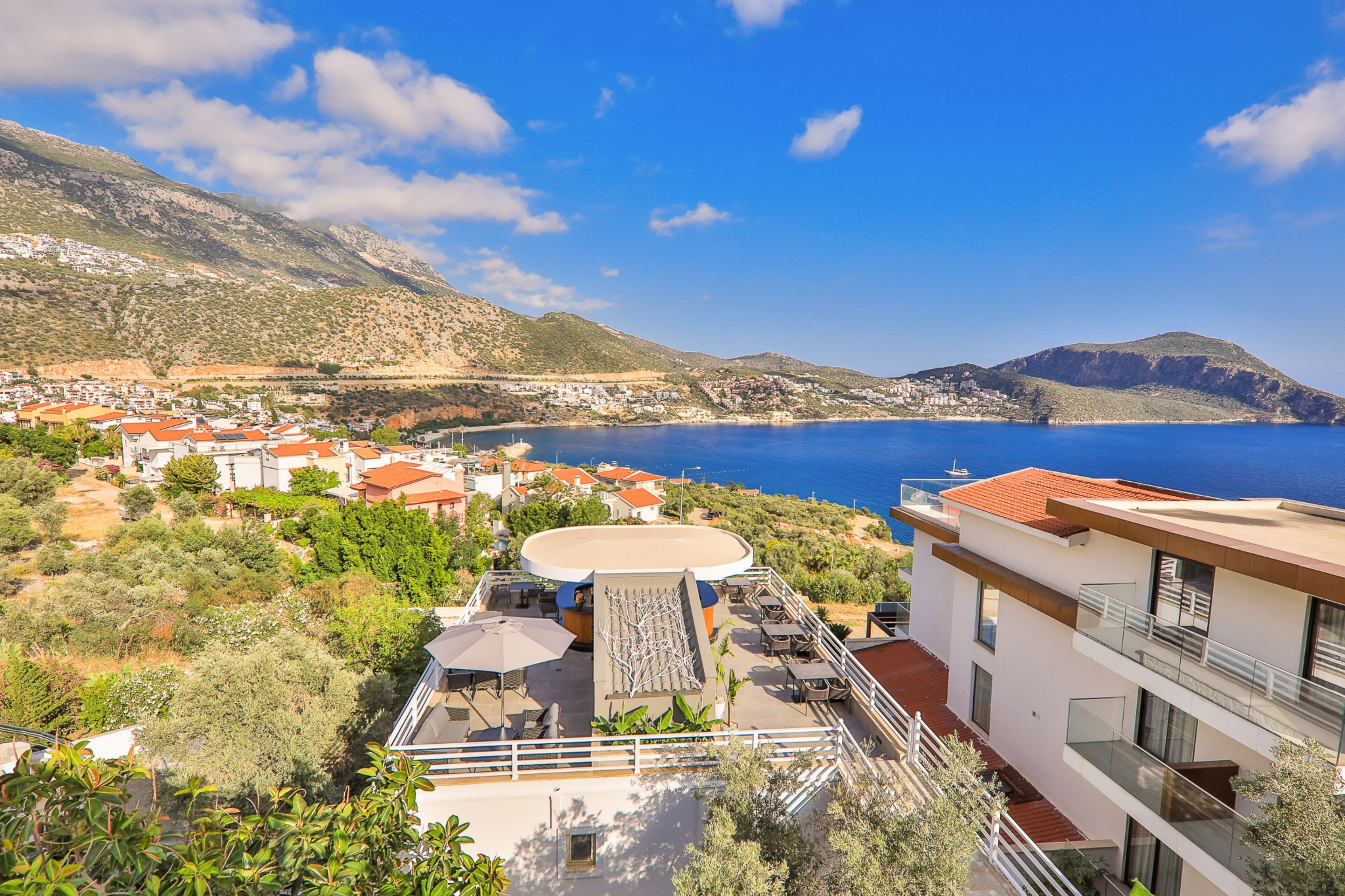 A coastal building with a terrace overlooking a bay, surrounded by greenery and hills.