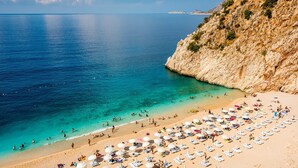 A beach with clear turquoise water, a rocky cliff, and numerous sun loungers and umbrellas. - Kalkan Saray Suites - Adults Only (Kas)