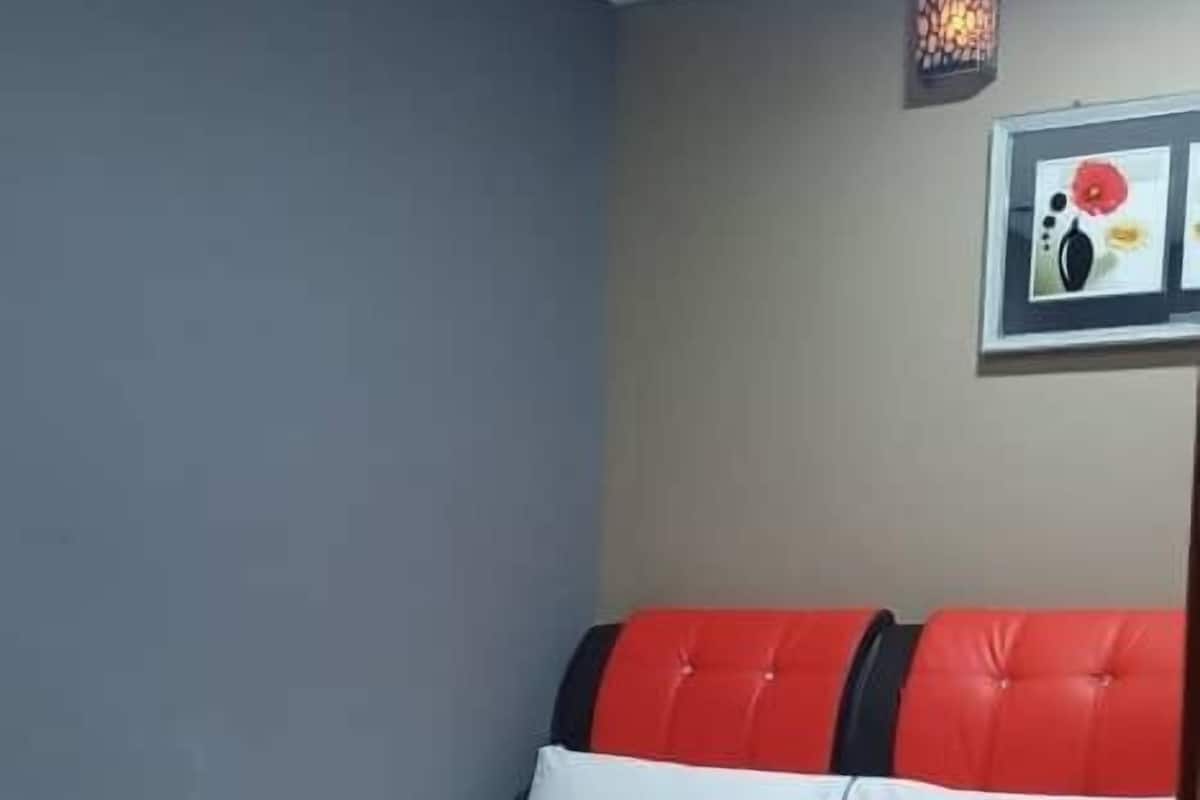Un lit bien fait, avec une tête de lit blanche et rouge, un tableau encadré au mur et un luminaire au plafond.