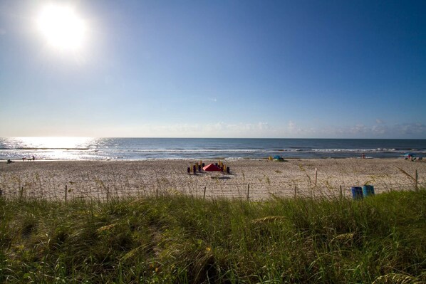 Una playa con cielo despejado, mar tranquilo y una carpa roja a lo lejos.