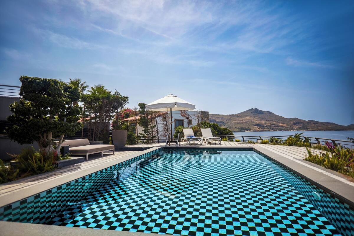 Piscina com padrão quadriculado, cadeiras de descanso, um guarda-sol e vista para montanha e lago.