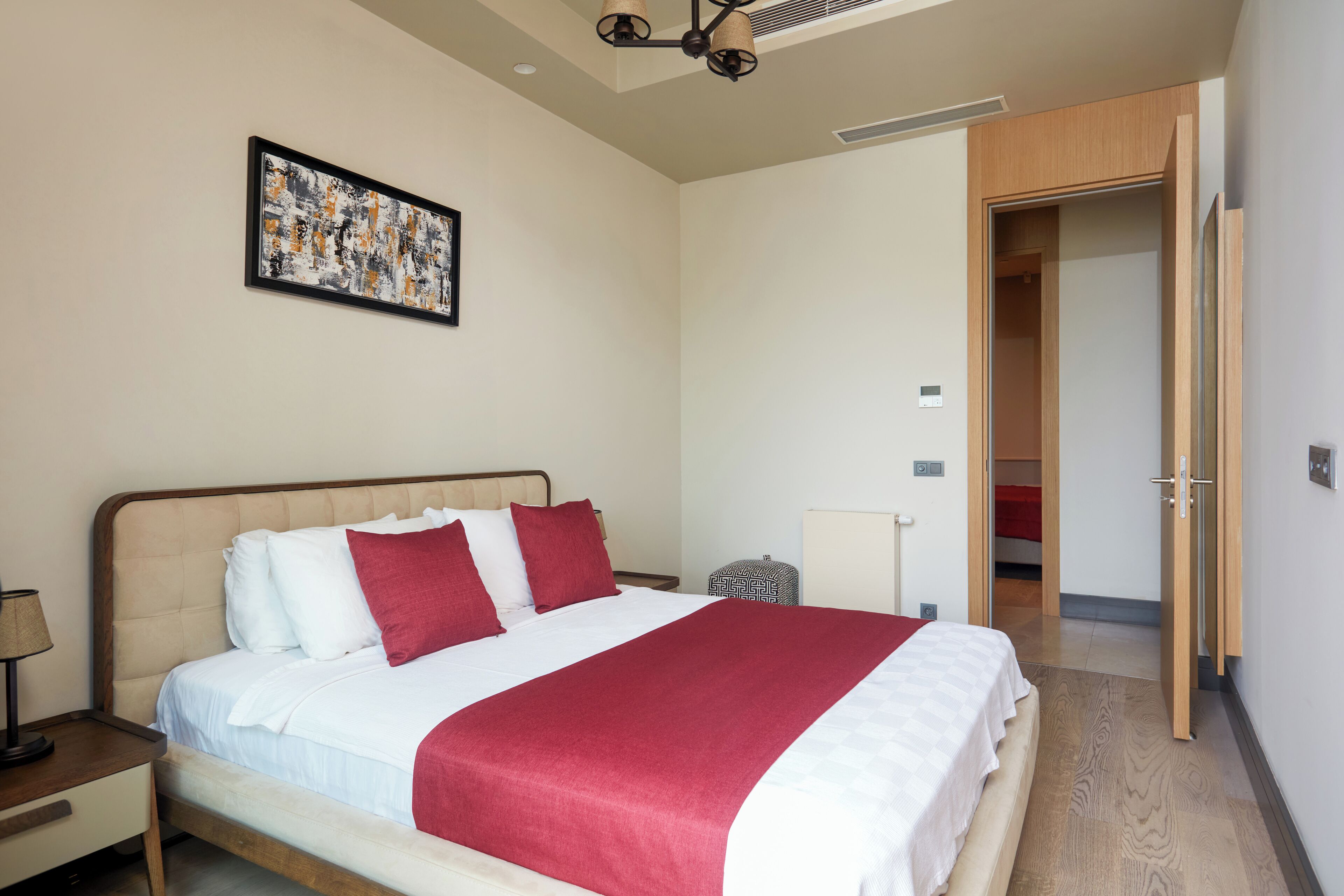 A neatly made bed with a red and white bedspread, a bedside table with a lamp, and a framed artwork on the wall.