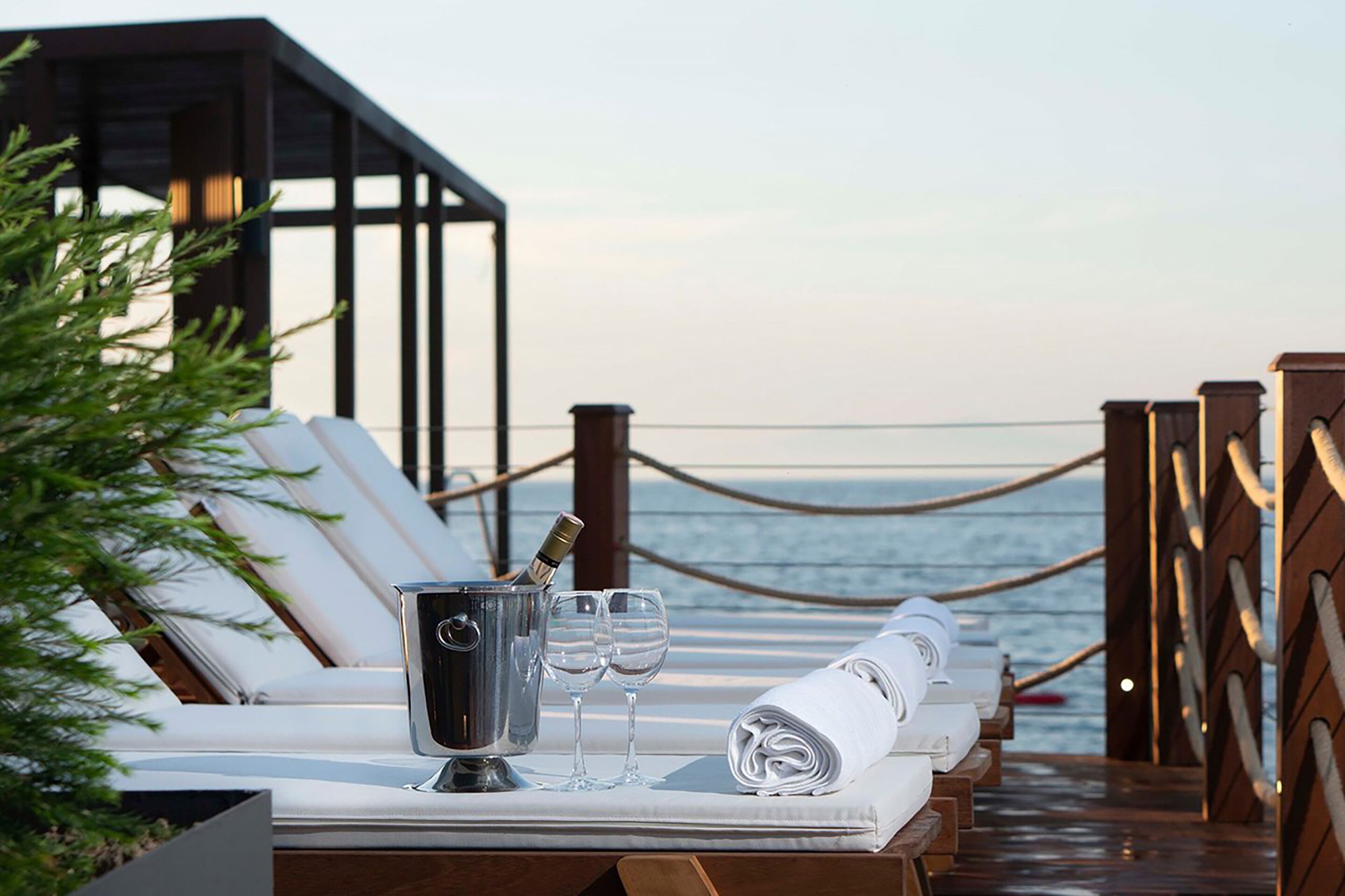 A sunlit outdoor lounge area with a view of the sea, featuring a table with a champagne bucket, glasses, and towels.