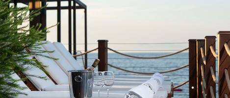 A sunlit outdoor lounge area with a view of the sea, featuring a table with a champagne bucket, glasses, and towels.