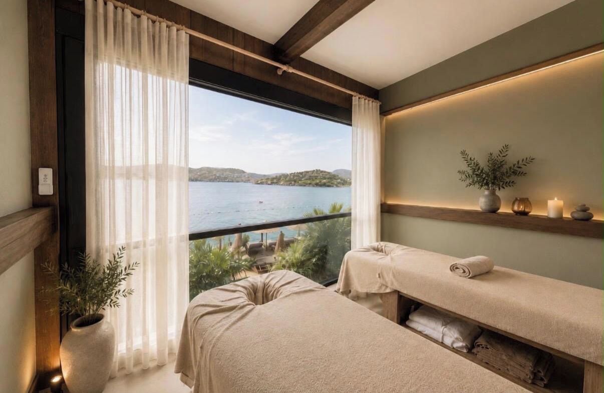 Una habitación con un ventanal grande, dos camillas de masaje, una estantería con velas y una planta, y vistas a un lago.