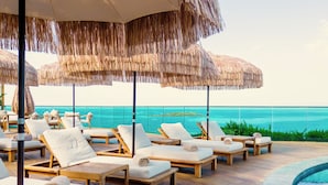 Une terrasse en bois avec des chaises longues et des parasols en chaume, donnant sur une piscine et l’océan.