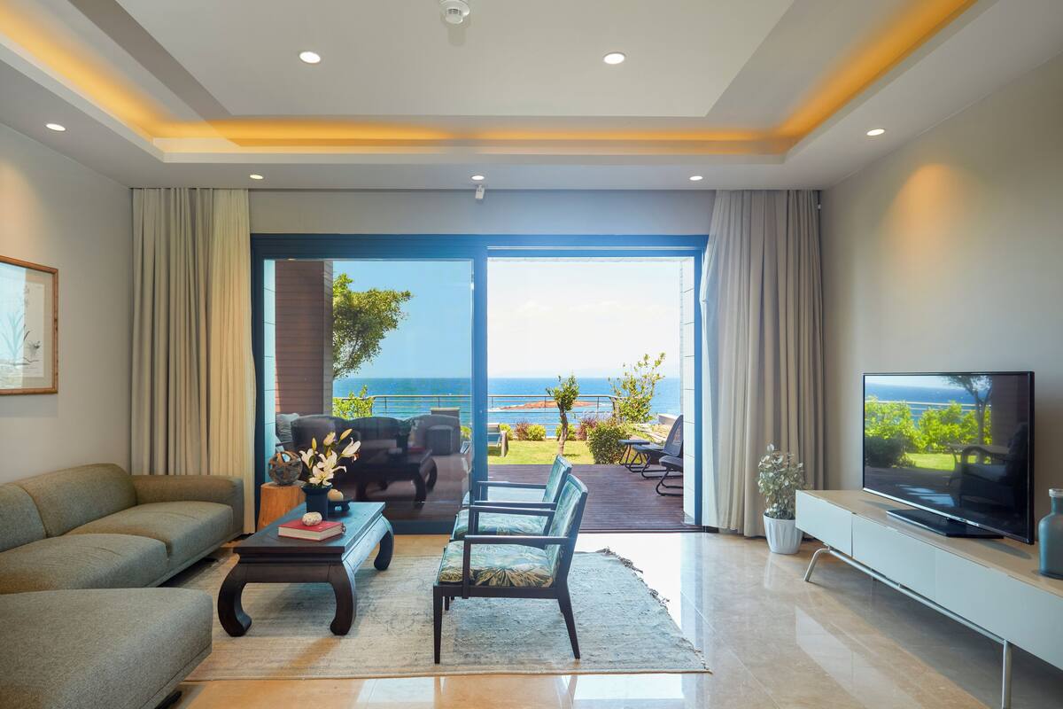 Uma sala de estar moderna com sofá, mesa de centro e televisão, com acesso a uma varanda e vista para o mar.