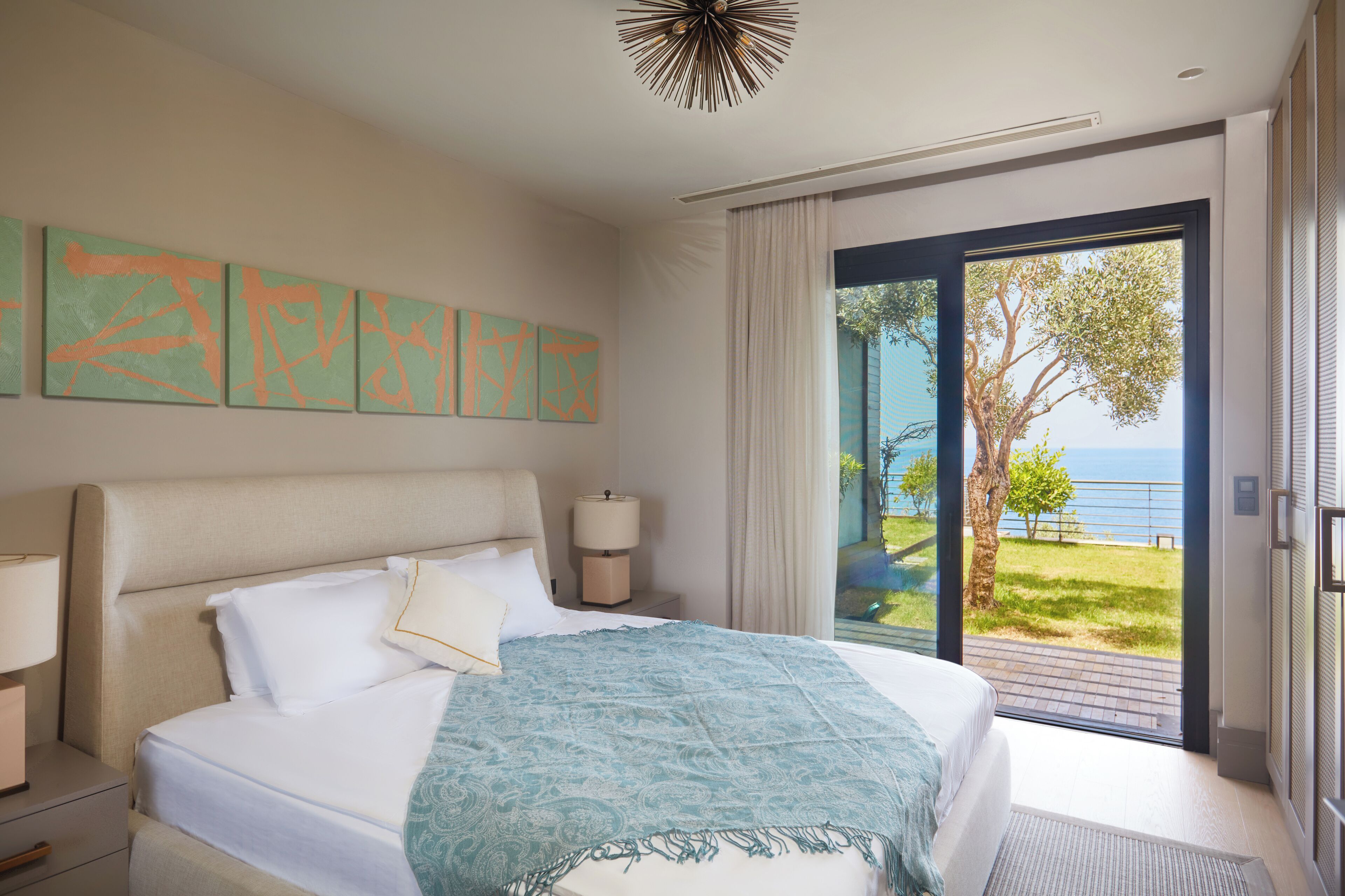 Une chambre d’hôtel avec un grand lit, une table de chevet, une lampe et une vue sur la mer, accessible par une porte-fenêtre coulissante.