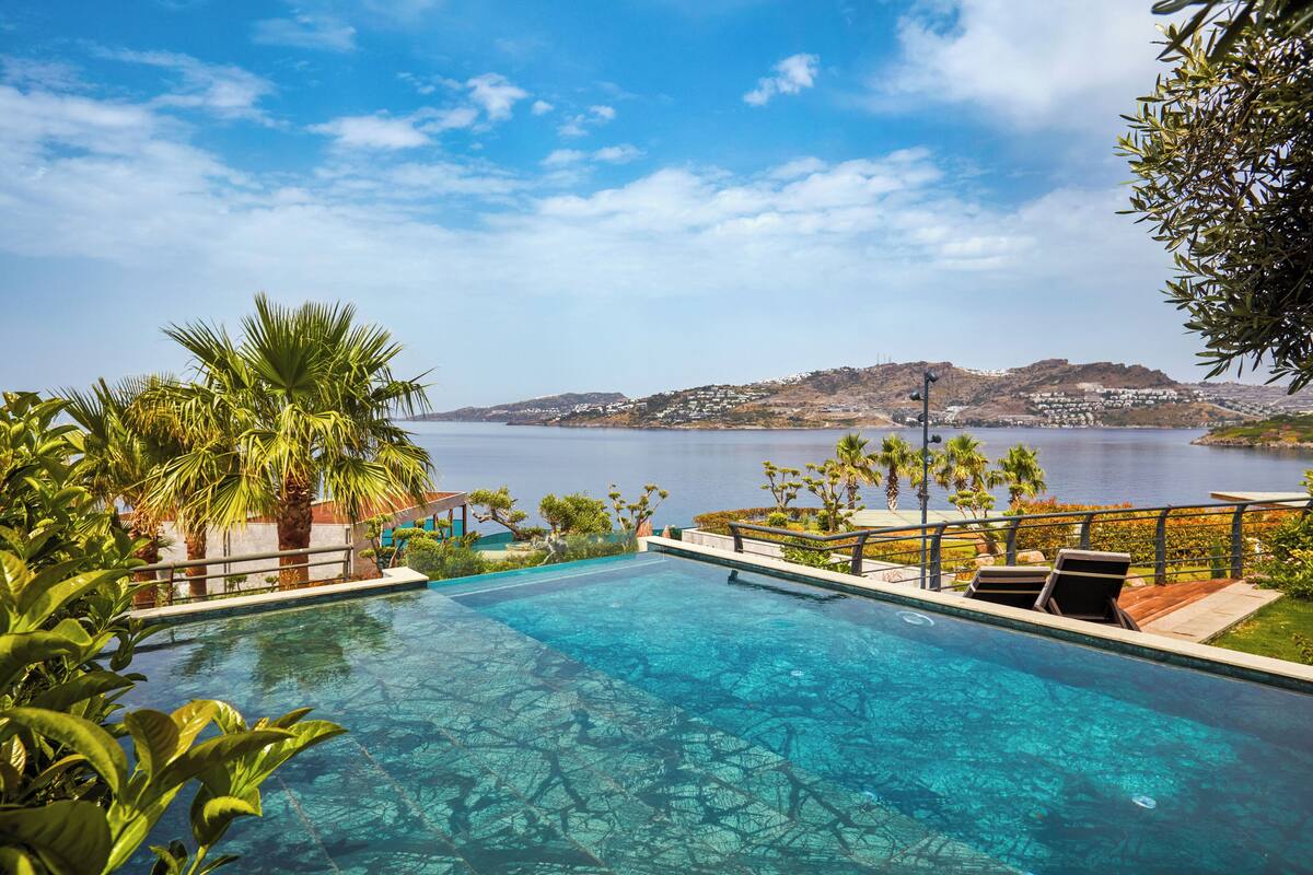 Piscina azul límpida com vista para o mar e colinas ao fundo.