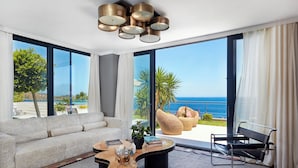 Un salon moderne avec un canapé, une table basse et un fauteuil, de grandes fenêtres offrant une vue sur l’océan et un balcon agrémenté de plantes.