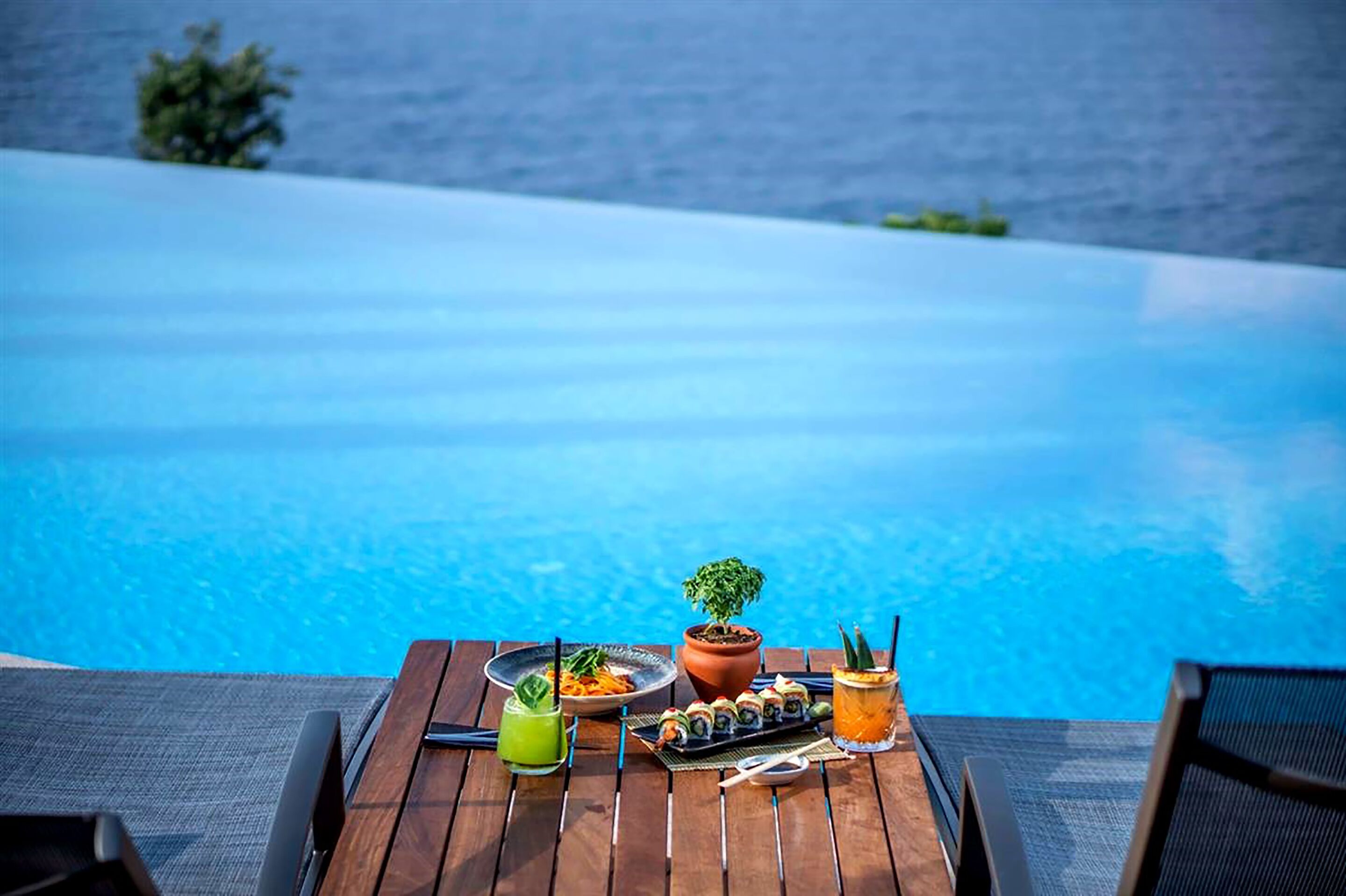 Mesa de madeira com refeição e bebidas à beira de uma piscina, com vista para um corpo d'água azul e límpido.