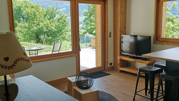 Une chambre moderne avec un plancher en bois, un téléviseur posé sur un meuble en bois, une table à manger avec des chaises et de grandes fenêtres offrant une vue sur la verdure et les montagnes.