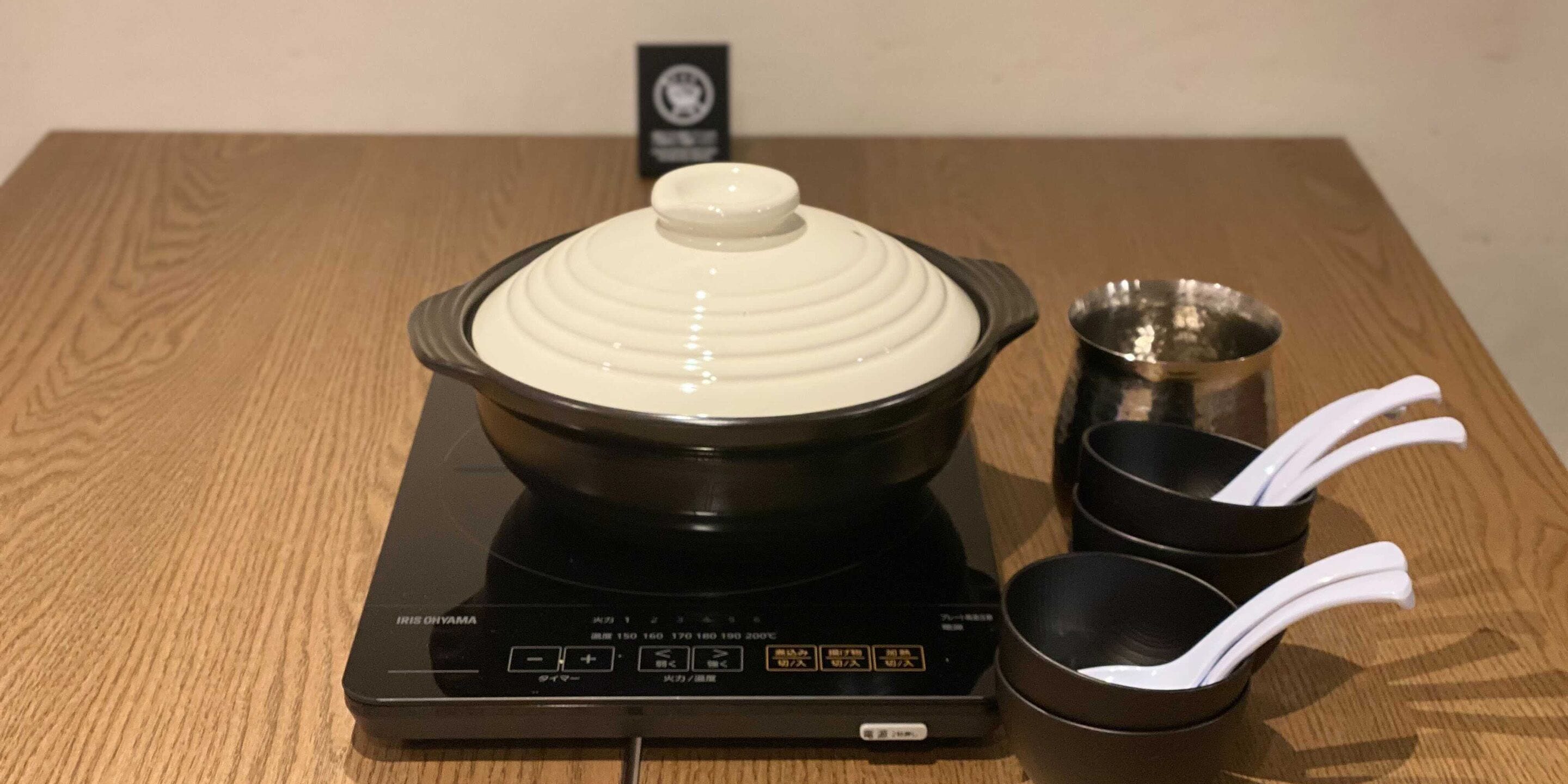 A black induction cooktop with a white pot on top, two black bowls, and a metal cup on a wooden table.