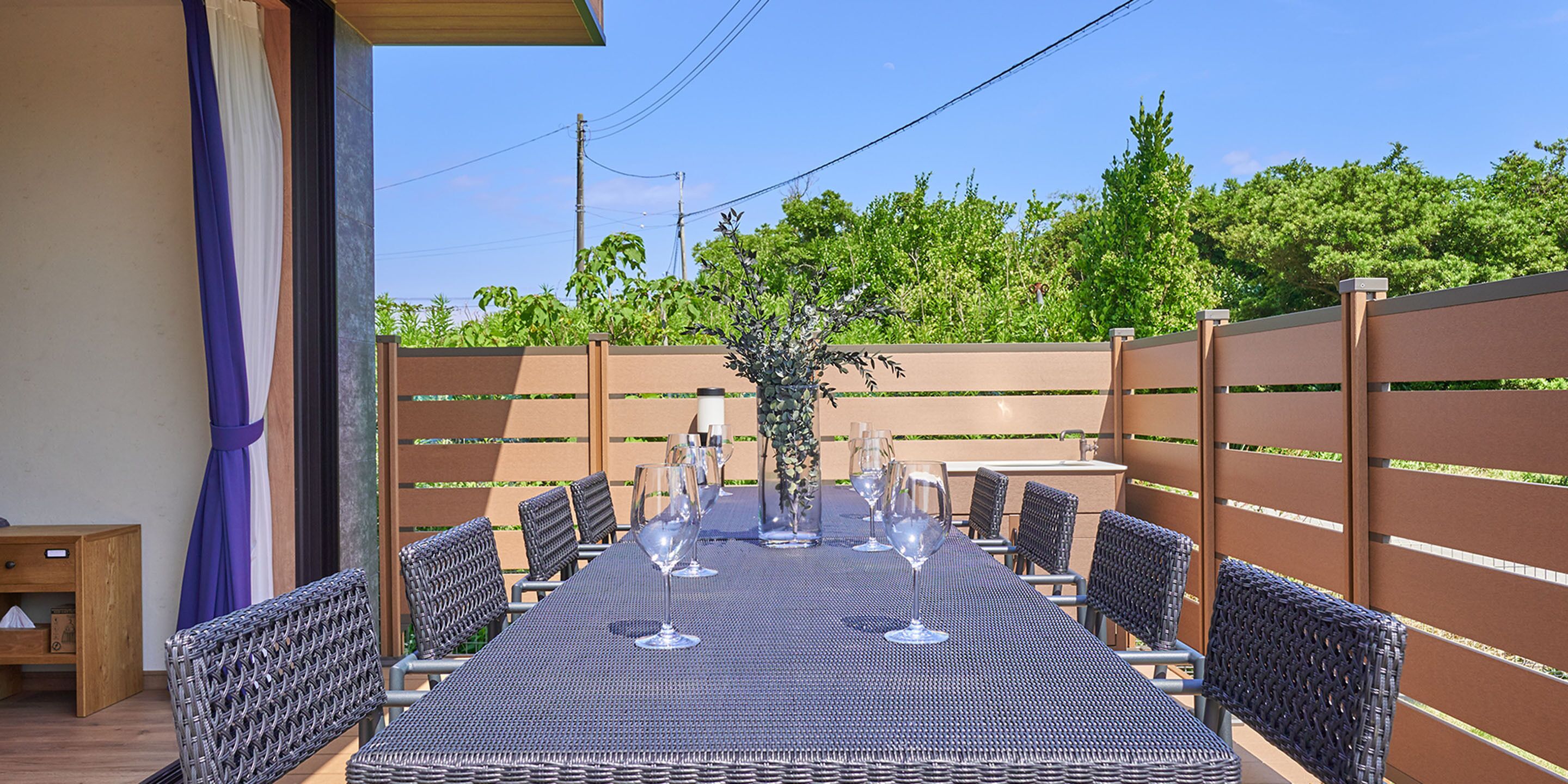 An outdoor dining area with a long table set for six, surrounded by wicker chairs, a wooden side table, and a decorative plant centerpiece.