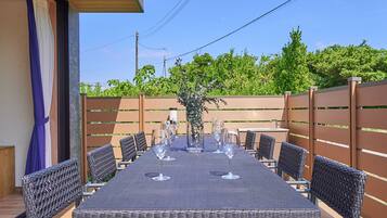 An outdoor dining area with a long table set for six, surrounded by wicker chairs, a wooden side table, and a decorative plant centerpiece.