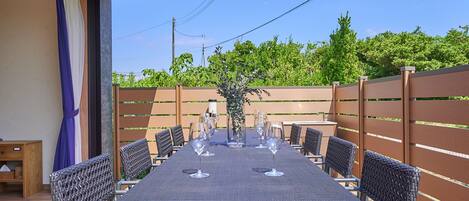 An outdoor dining area with a long table set for six, surrounded by wicker chairs, a wooden side table, and a decorative plant centerpiece.