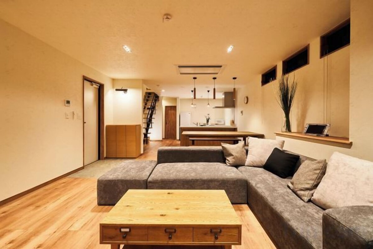 Una sala de estar moderna con un sofá seccional, una mesa de centro de madera y una cocina al fondo.