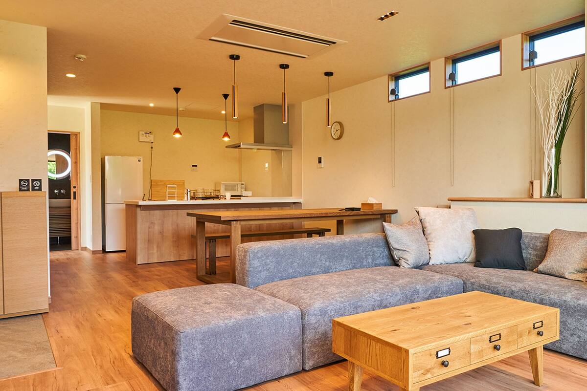 A modern living room with a grey sectional sofa, a wooden coffee table, and a kitchen area in the background.