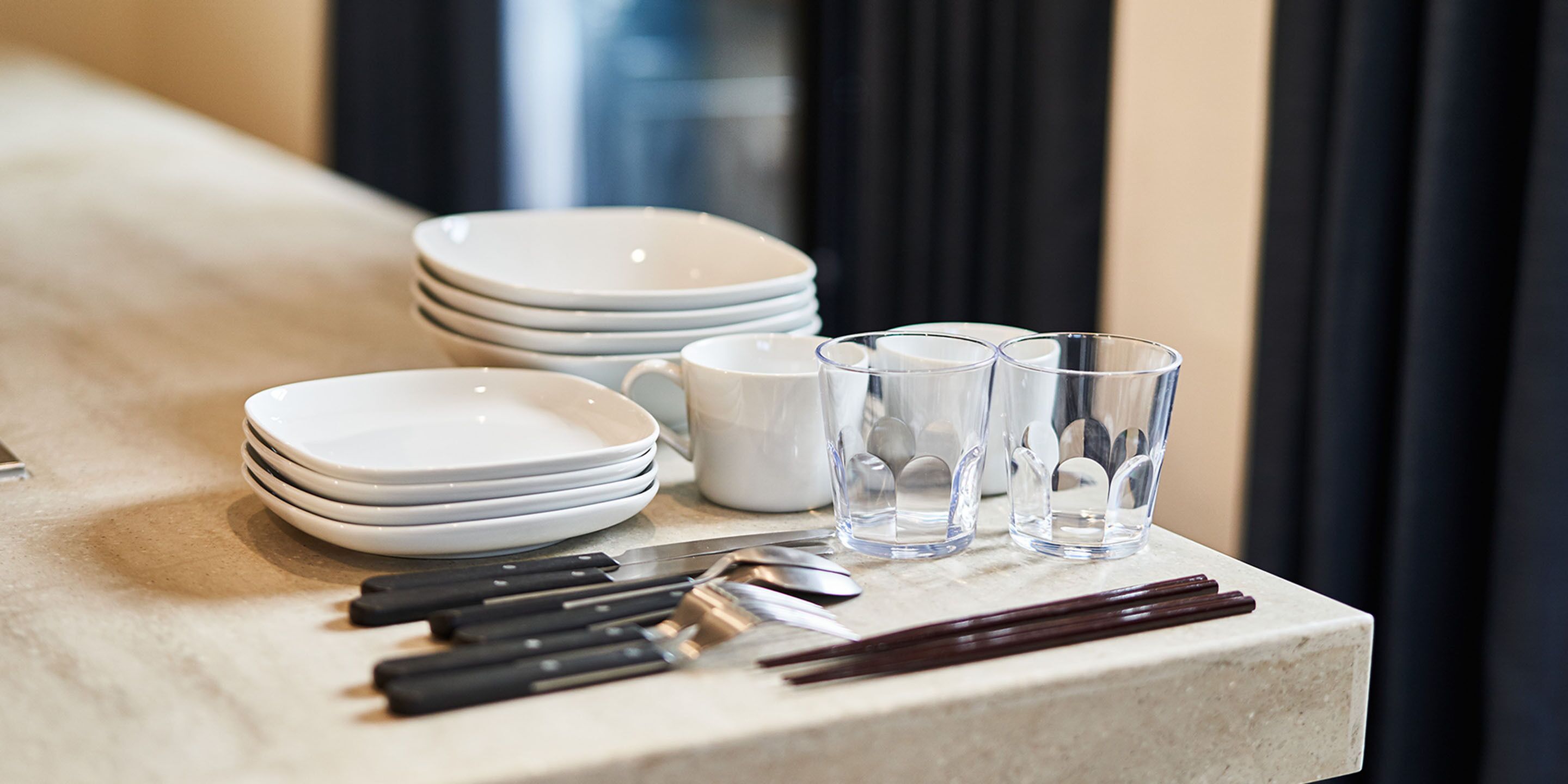 A table with stacked white plates, clear glasses, black-handled knives, and a pair of chopsticks.