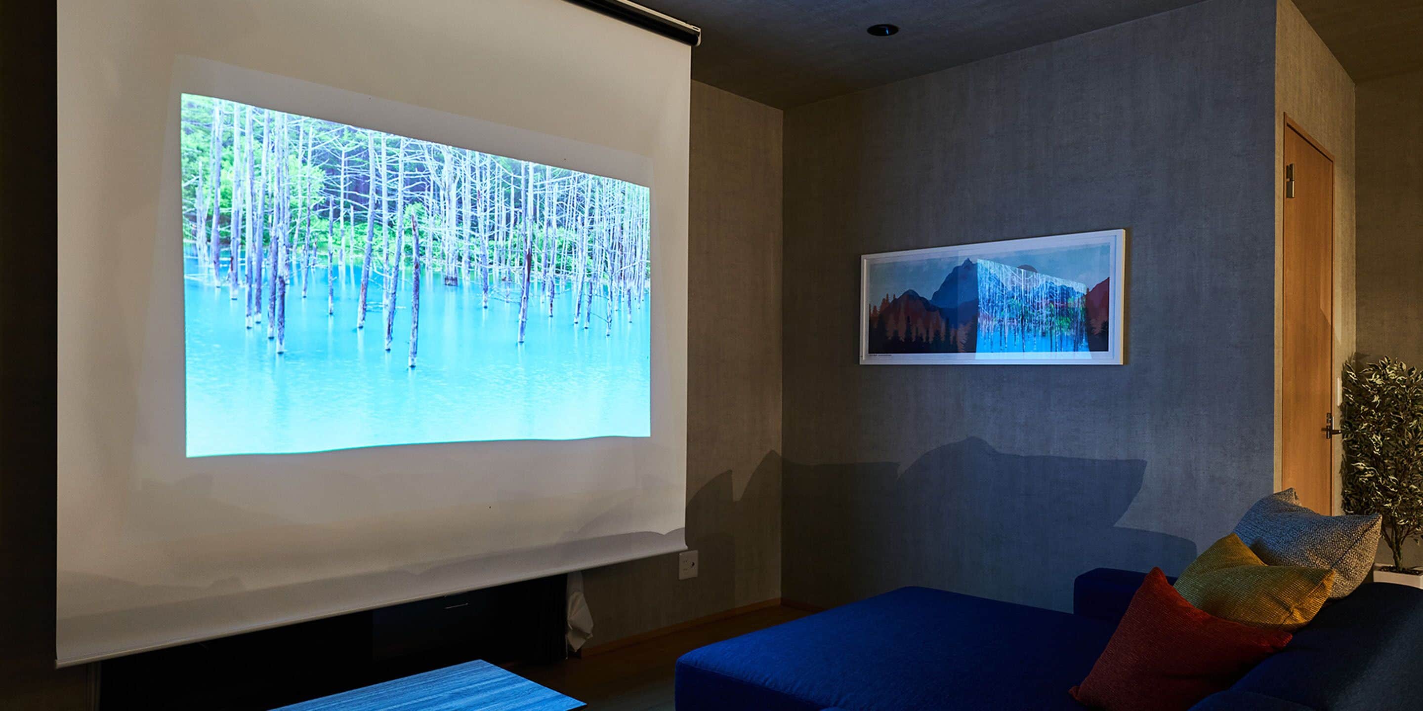 Uma tela de projeção exibindo uma paisagem natural, um quadro na parede e um sofá azul com almofadas coloridas.