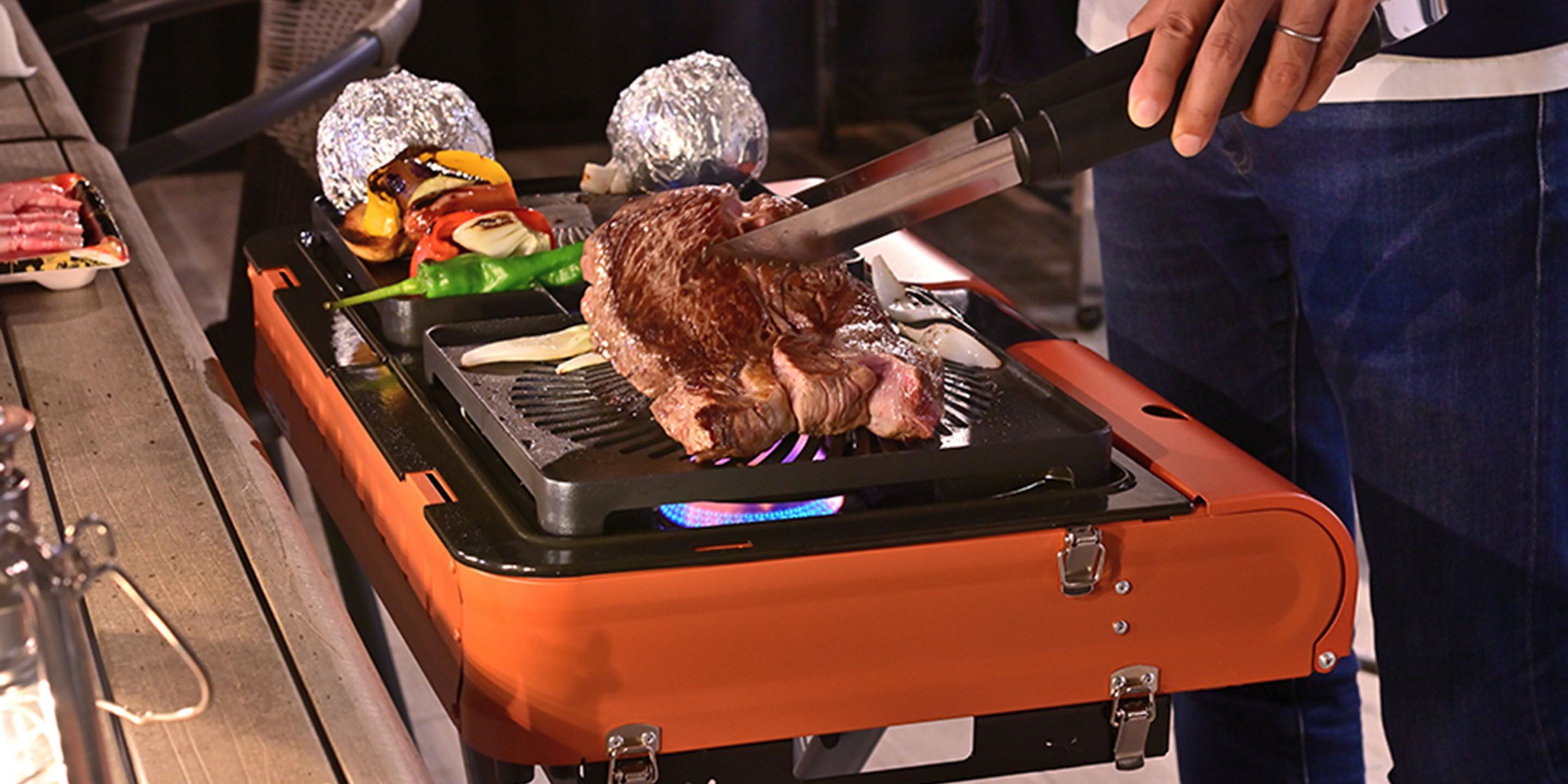 Un barbecue portable sur lequel on fait griller un steak, entouré de légumes et d’aliments emballés dans du papier d’aluminium.