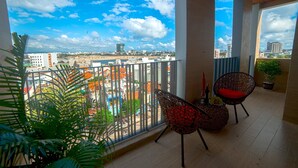 Un balcón con vistas a un paisaje urbano, una silla de mimbre, una mesita con una botella y uvas, y una planta en maceta.