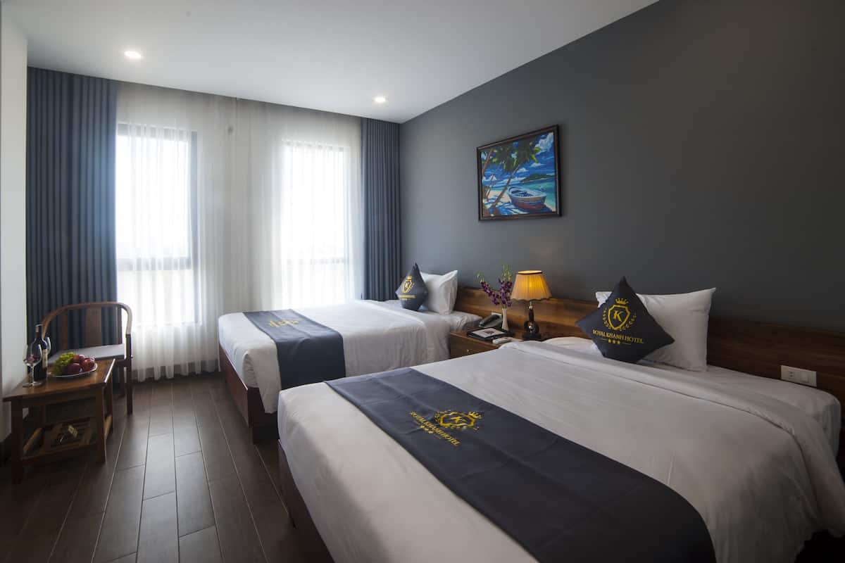 Une chambre d’hôtel avec deux lits, une table en bois sur laquelle se trouve un panier de fruits, une chaise, une table de chevet avec une lampe et un tableau accroché au mur.