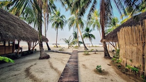A wooden pathway leading to a beach with palm trees and thatched-roof huts.