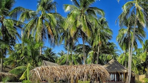 A beach with two lounge chairs and a thatched umbrella, surrounded by palm trees and a stone wall.